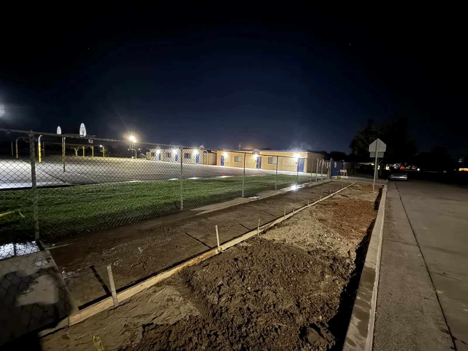 Trench and forms along a playground fence, lit by work lights before dawn